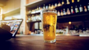 A close-up of a frothy, cold pint of golden beer in a transparent glass, set on a wooden table in a dimly lit pub, evoking a vintage, celebratory atmosphere.