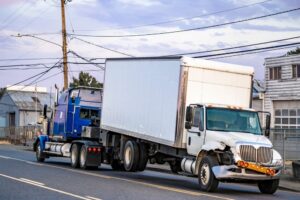 Mobile blue roadside assistance powerful tow big rig semi truck tractor towing broken after accident day cab semi truck with box trailer driving on the local road at evening time.