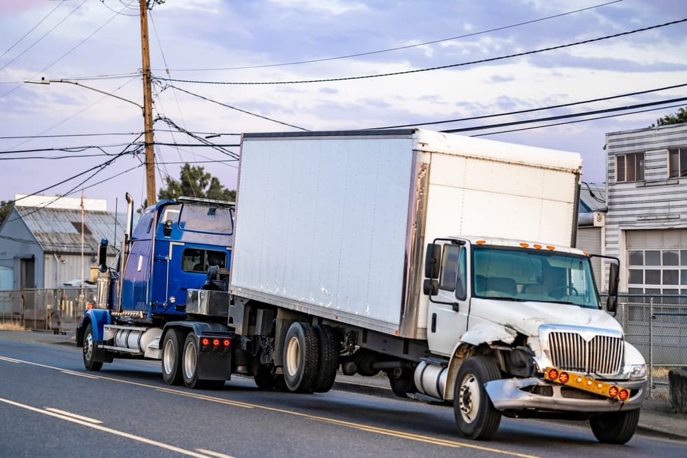 Mobile blue roadside assistance powerful tow big rig semi truck tractor towing broken after accident day cab semi truck with box trailer driving on the local road at evening time.