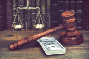 Judge Gavel, Scale Of Justice With US Dollars And Law Book On Judges Table In Courtroom. 
